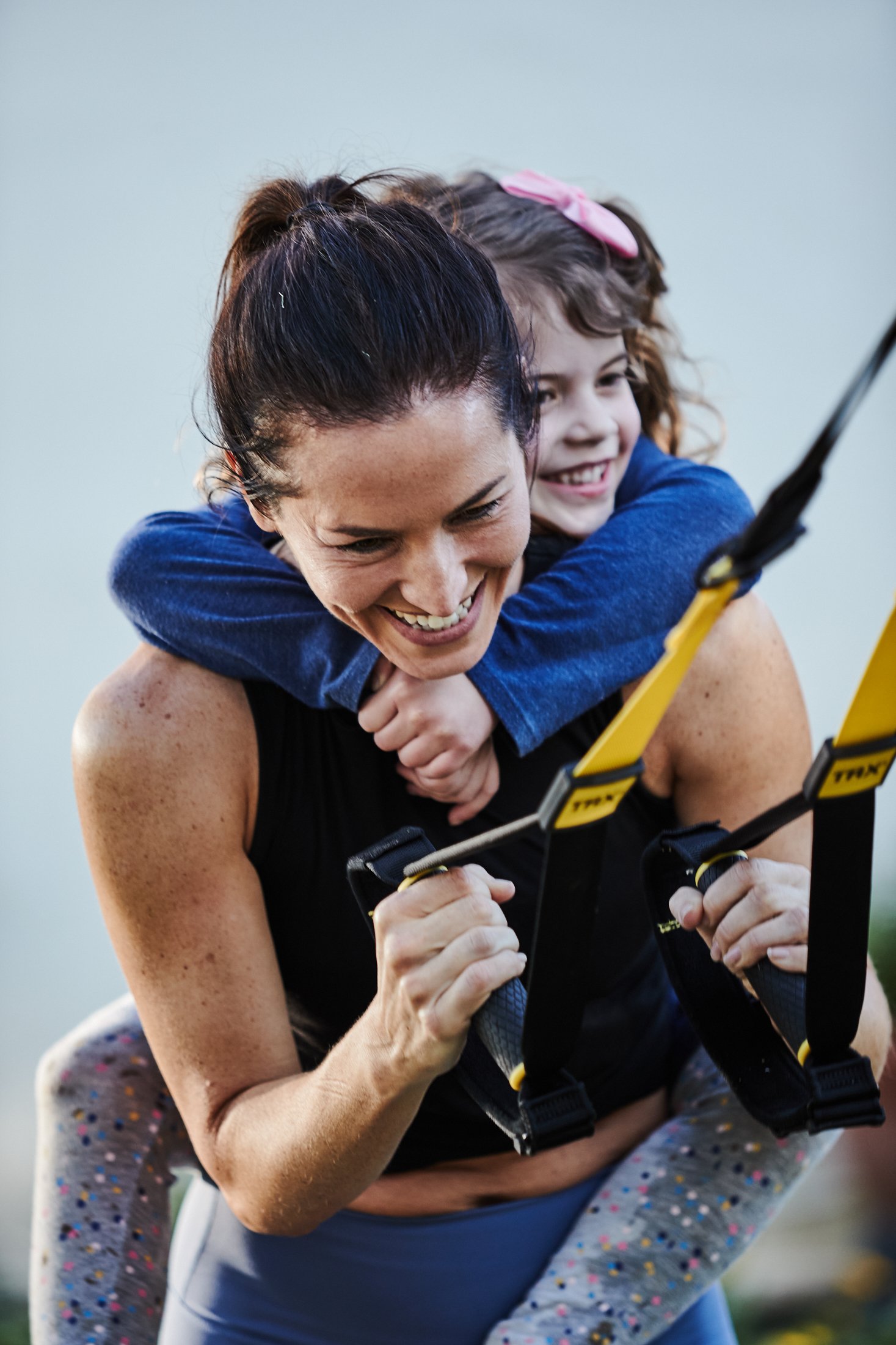Kristin Leffel performs a TRX squat with one of her children hanging on her back