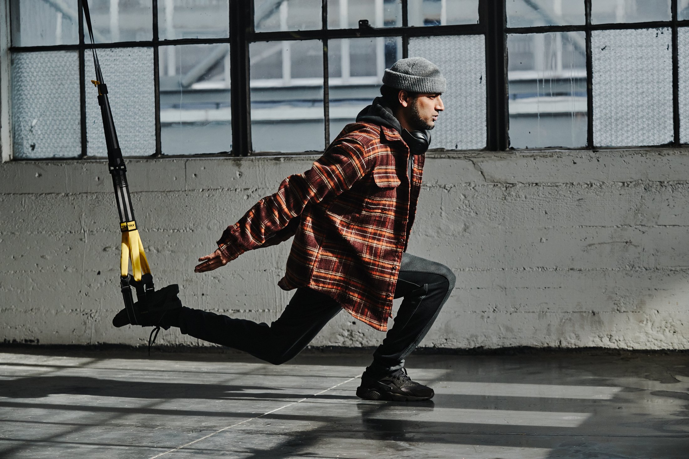 TJ Yale, wearing a beanie, red flannel shirt, and black pants and sneakers, performs a TRX Lunge.
