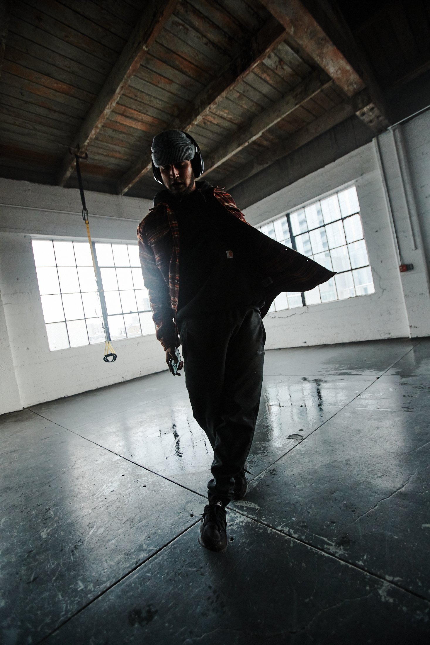 TJ Yale, wearing a beanie, flannel shirt, pants, and sneakers, performs a dance move in an industrial space with windows behind him.
