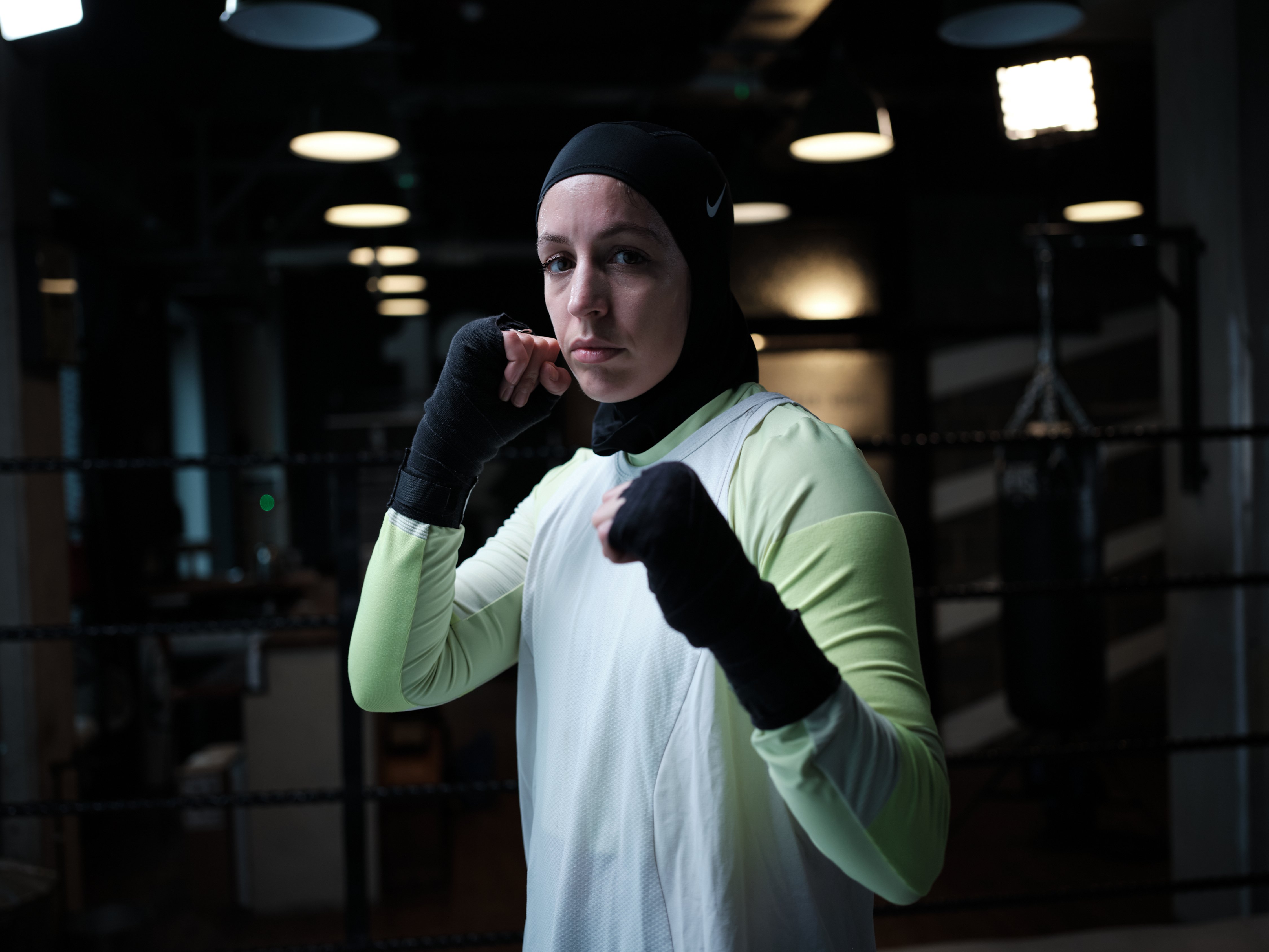 Nesrine Dally in a fighting position. She wears a black hijab, black handraps, white vest, and longsleeve green shirt. She is in a boxing gym.