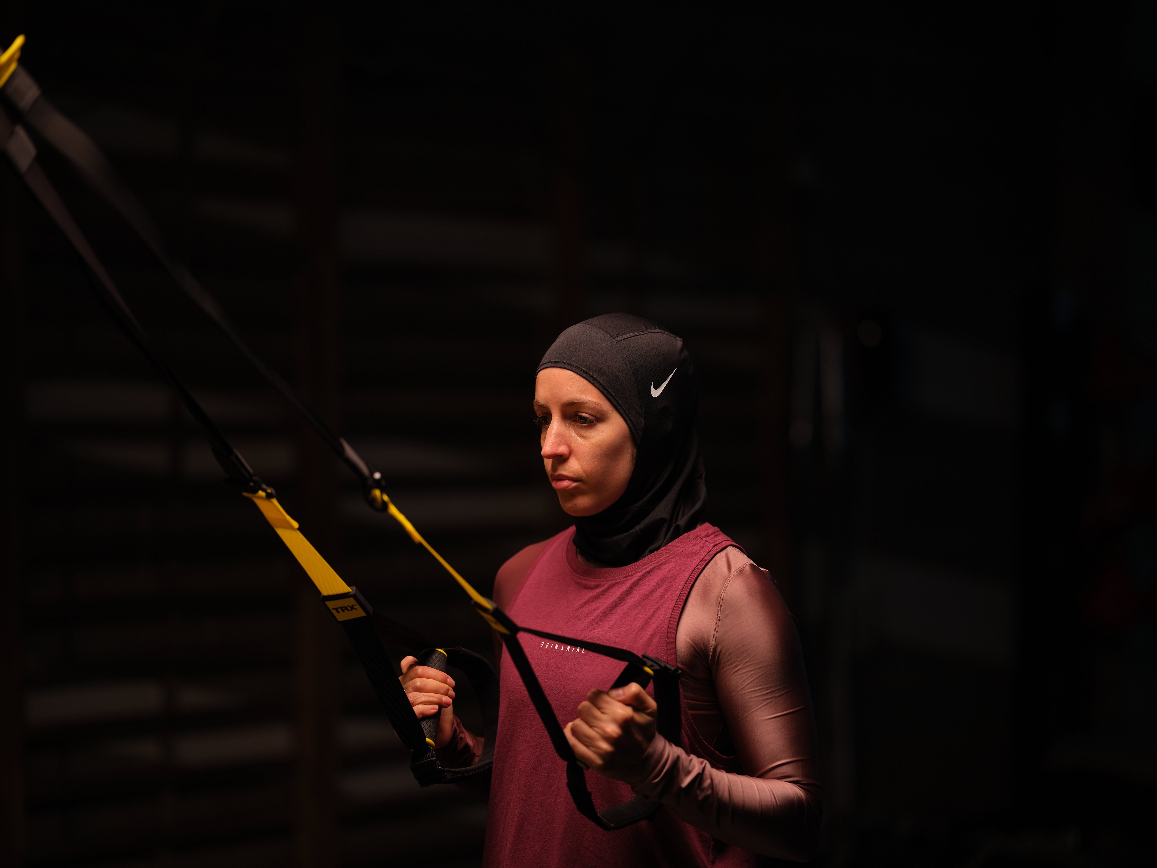 Nesrine Dally using the TRX Suspension Trainer for a TRX Row. She wears a black hijab, pink longsleeve shirt, and dark rose vest.