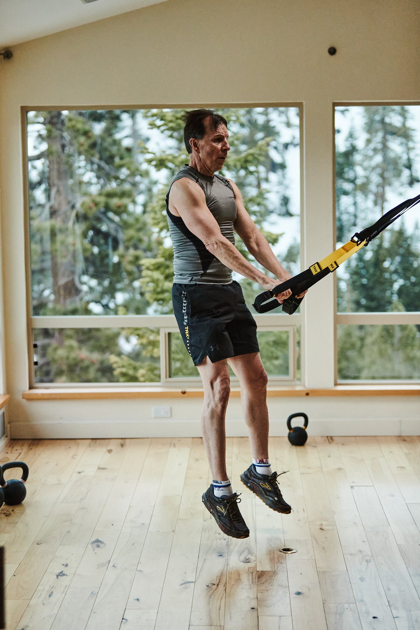 Walt Raineri, wearing a grey sleevess top, black shorts, and black sneakers, does a TRX squat jump. He's in a large room with windows overlooking snowcapped trees.
